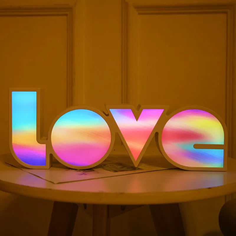Rainbow-coloured LED LOVE decorative lamp with plastic shell on a table.