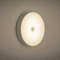 Small white circular motion sensing LED night light with USB charging cable and adhesive tape.