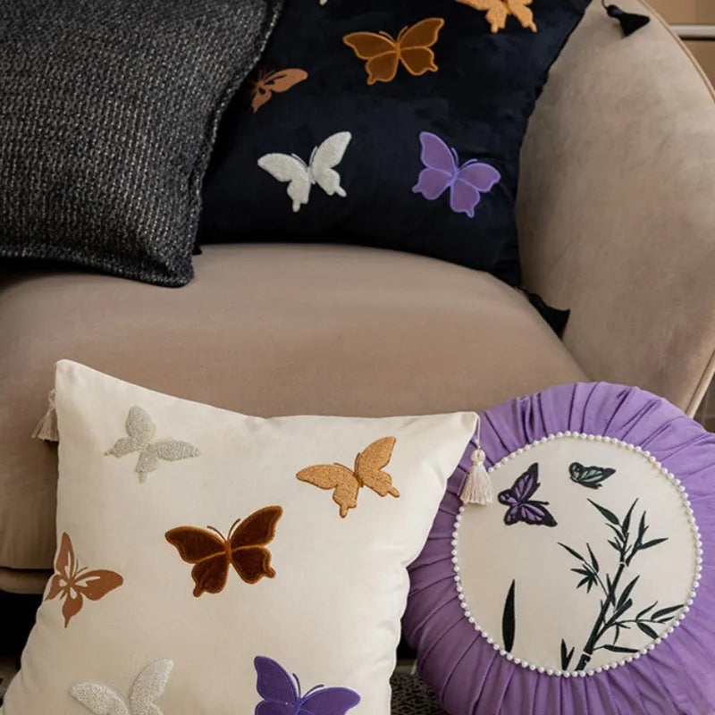 Square beige cushion with butterfly embroidery on a sofa, showing purple and black colour options.