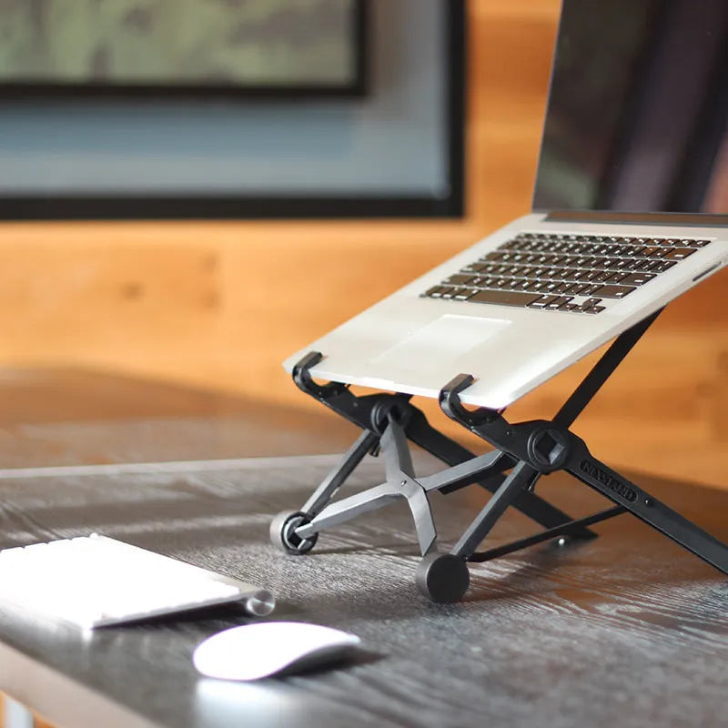 Black folding portable laptop stand with reinforced nylon construction shown unfolded beside laptop