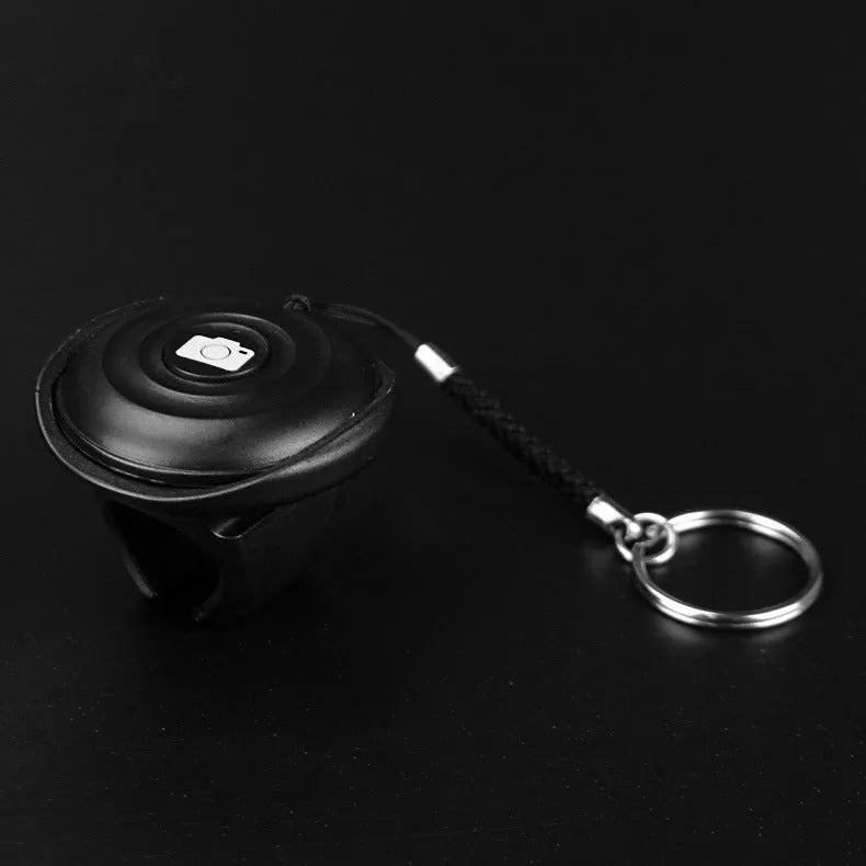 Black Bluetooth remote shutter for smartphones on a white background, shown next to a mobile phone.