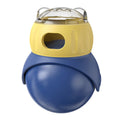 Yellow and blue owl-shaped plastic tumbler toy for cats, a slow-feeder puzzle on a wooden floor.