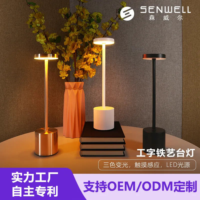 Modern minimalist iron table lamp with silver finish, I-shaped silhouette, emitting warm ambient light on a bedside table