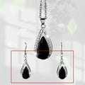 Close-up of a black alloy teardrop pendant necklace and matching drop earrings with glass details on a plain background.