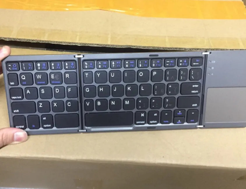 Black tri-fold folding Bluetooth keyboard with touchpad shown open and closed, next to a mobile phone.