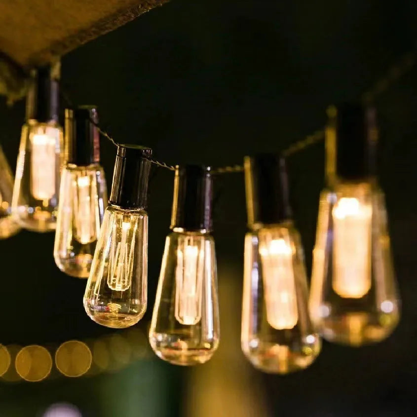 Retro long bulb outdoor string lights in a garden setting, showing solar panel, battery box, and USB power options.