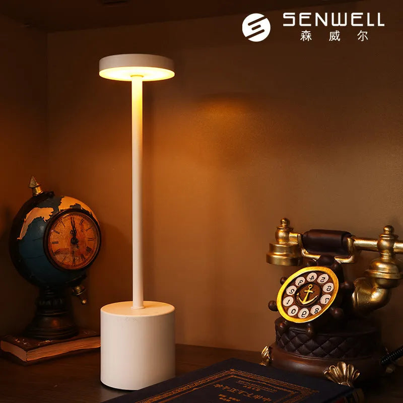 Modern minimalist iron table lamp with silver finish, I-shaped silhouette, emitting warm ambient light on a bedside table