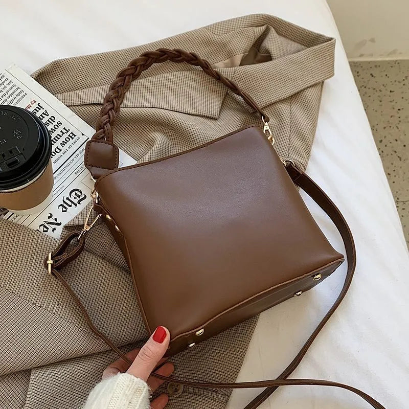 Khaki woven texture one-shoulder crossbody bucket bag with an underarm strap on a table.