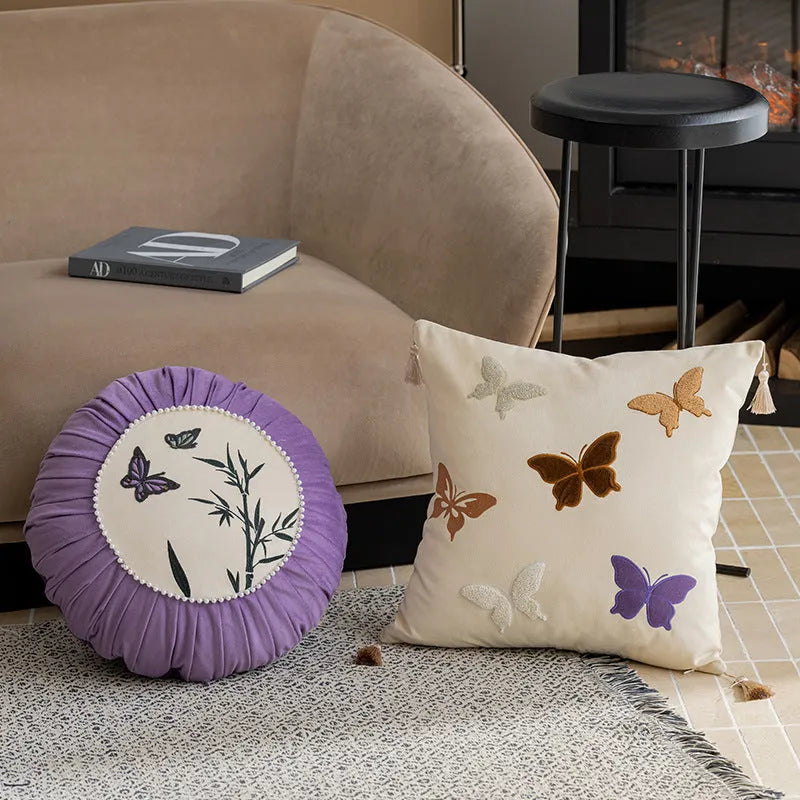 Square beige cushion with butterfly embroidery on a sofa, showing purple and black colour options.