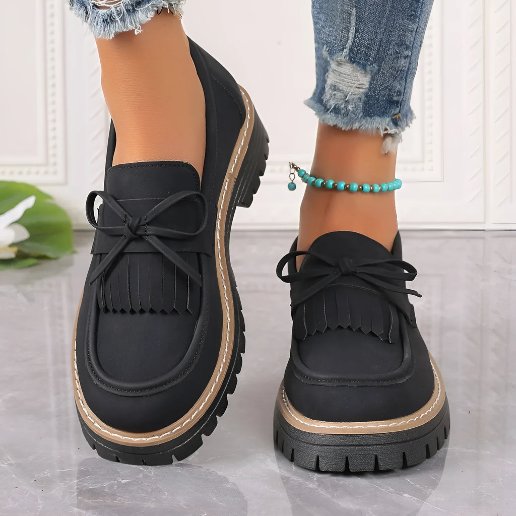 A pair of black plus size women's loafers with a tassel and bowknot decoration on a chunky rubber sole.