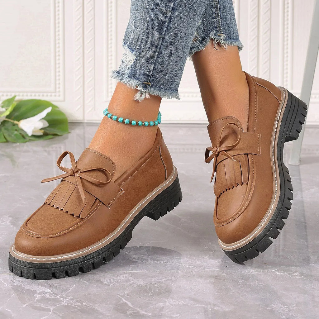 A pair of black plus size women's loafers with a tassel and bowknot decoration on a chunky rubber sole.