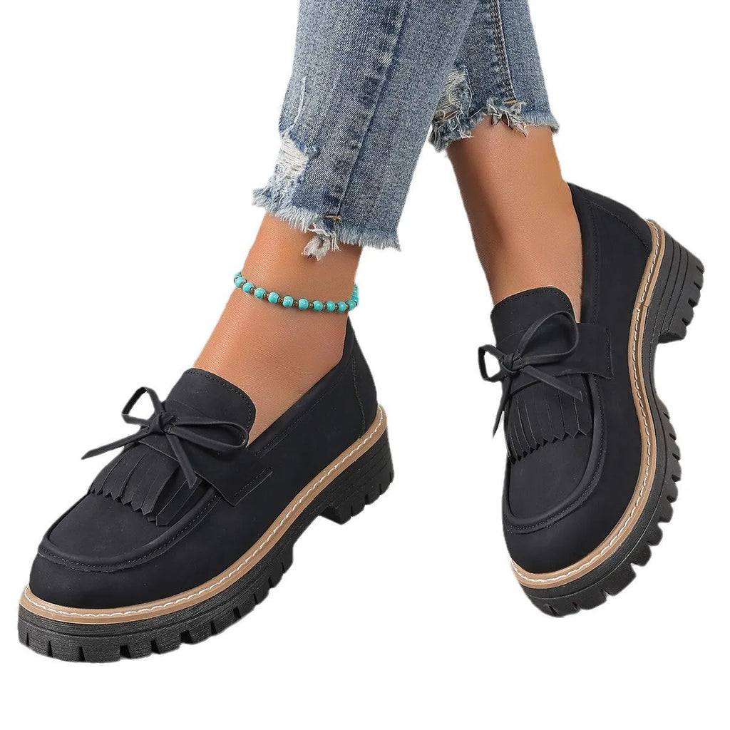 A pair of black plus size women's loafers with a tassel and bowknot decoration on a chunky rubber sole.