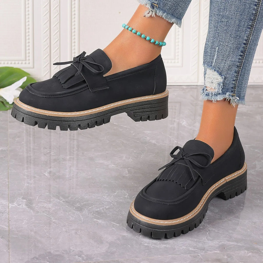 A pair of black plus size women's loafers with a tassel and bowknot decoration on a chunky rubber sole.