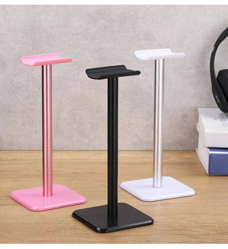 Detachable aluminum alloy headset hanger with TPU rubber cradle, shown in white, pink, and black, fixed to a table.