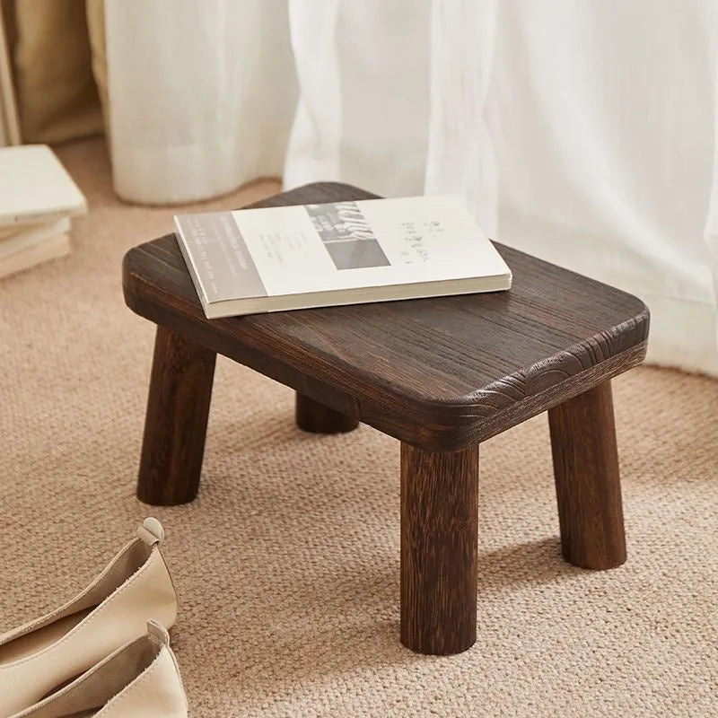 A solid wood stool with a distressed finish, used for changing shoes in a living room.