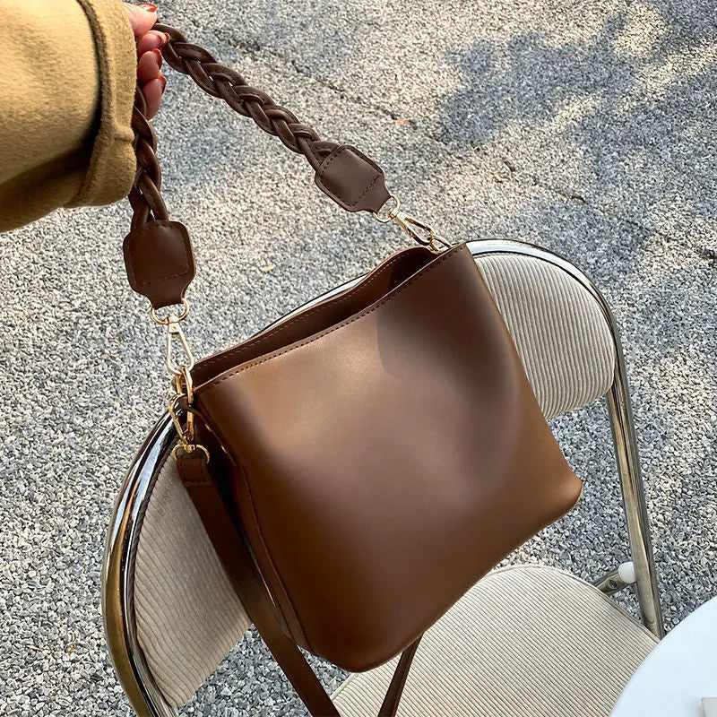 Khaki woven texture one-shoulder crossbody bucket bag with an underarm strap on a table.