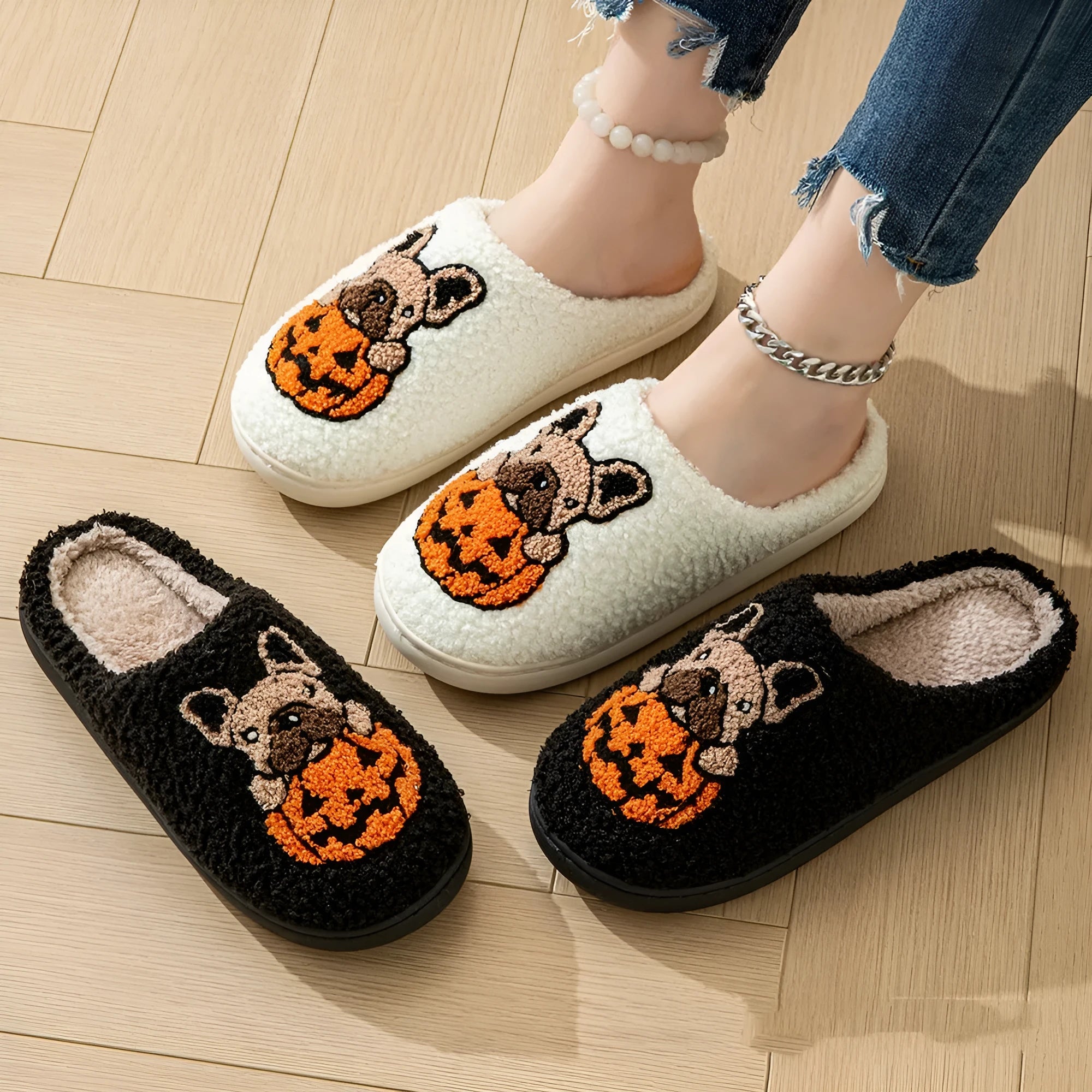 Pair of black plush Halloween slippers with pumpkin design on a wooden floor, showing round toe and textured TPR sole.