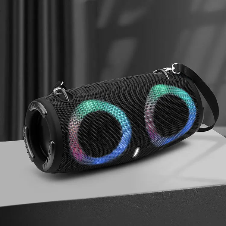 Cylindrical War Drum Bluetooth speaker with glowing RGB lights, a black metal grille, and a carrying strap on a wooden table.
