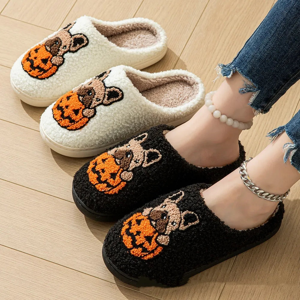 Pair of black plush Halloween slippers with pumpkin design on a wooden floor, showing round toe and textured TPR sole.