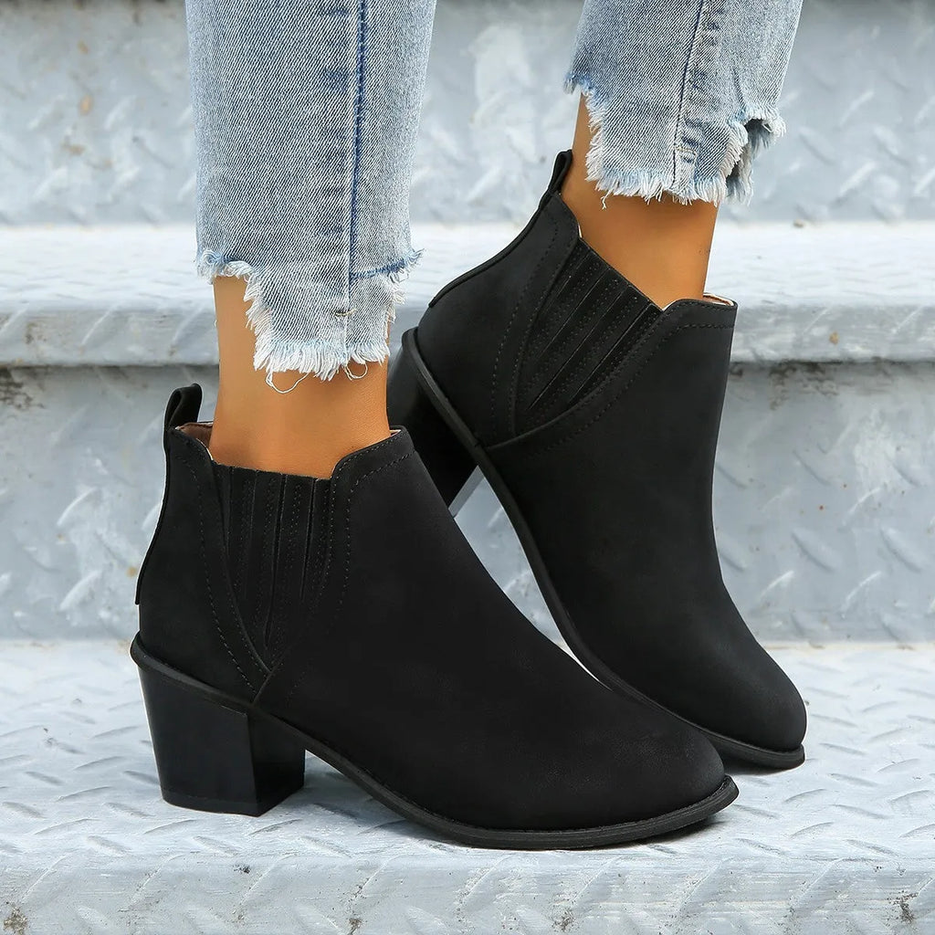 Women's black slip-on ankle boots with pointed toe and thick square heel on a wooden floor.