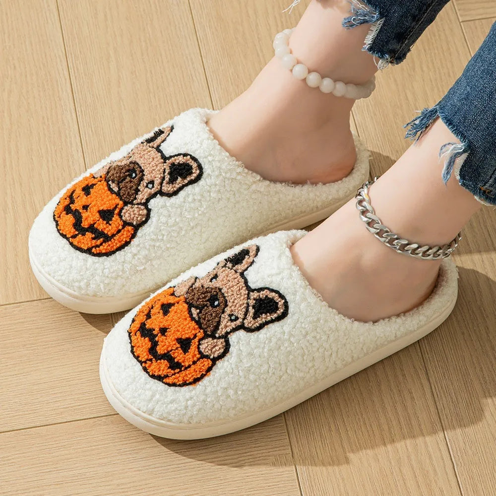 Pair of black plush Halloween slippers with pumpkin design on a wooden floor, showing round toe and textured TPR sole.