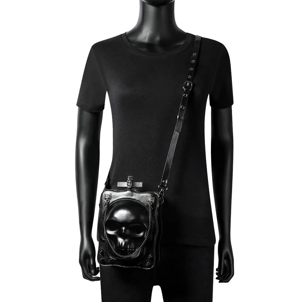Black vertical square crossbody bag with embossed skull design, chain strap, and metal rivets.