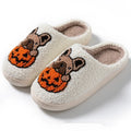 Pair of black plush Halloween slippers with pumpkin design on a wooden floor, showing round toe and textured TPR sole.