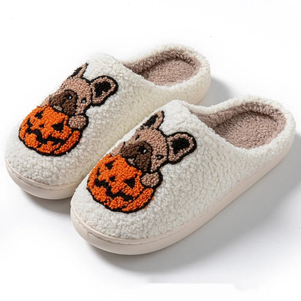 Pair of black plush Halloween slippers with pumpkin design on a wooden floor, showing round toe and textured TPR sole.