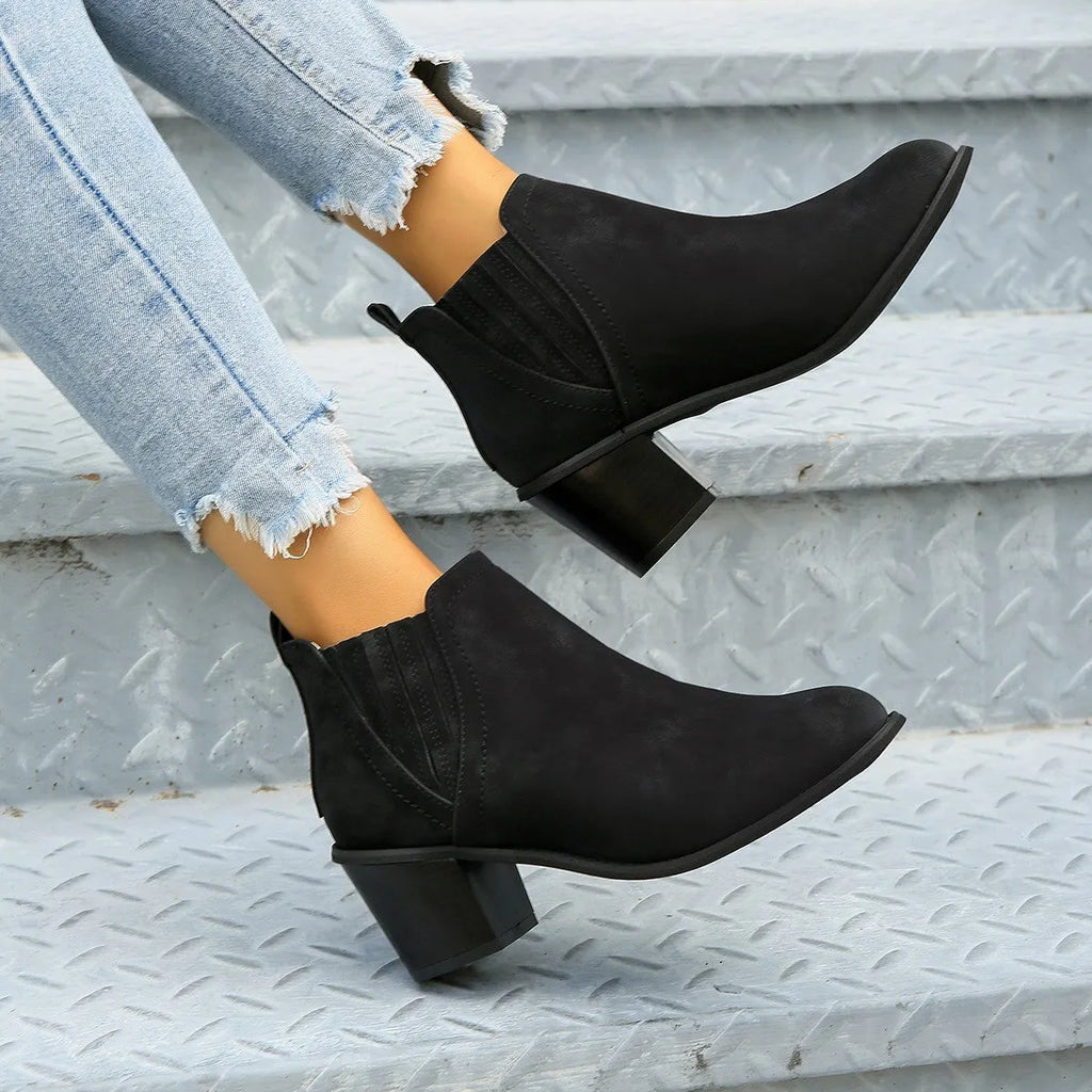 Women's black slip-on ankle boots with pointed toe and thick square heel on a wooden floor.